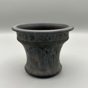Peut inclure: Un pot de fleur en céramique gris foncé avec une forme évasée en sablier. Le pot a un large bord plat et une surface texturée avec des stries de gris et de brun plus clairs. L'intérieur est d'une nuance de gris plus foncée.