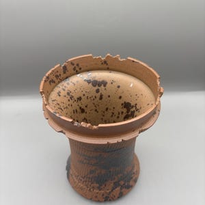 May include: A handmade ceramic vase with a unique, textured design. The vase features a cylindrical shape with a wide, slightly irregular rim. The exterior is a warm brown with dark brown speckles and patterns, giving it a rustic appearance.