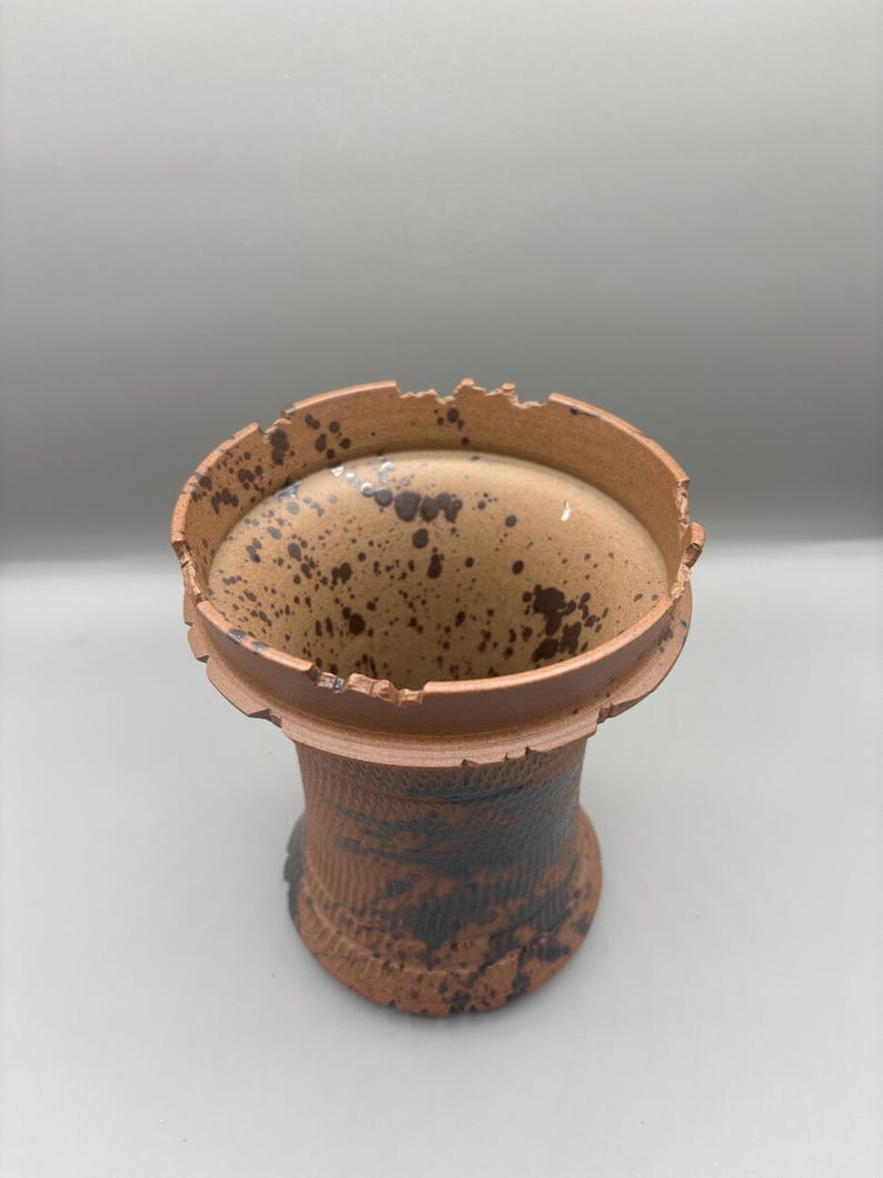 May include: A handmade ceramic vase with a unique, textured design. The vase features a cylindrical shape with a wide, slightly irregular rim. The exterior is a warm brown with dark brown speckles and patterns, giving it a rustic appearance.