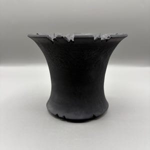 May include: A matte black vessel with a flared, wide rim and a narrow base. The exterior surface features a subtle, textured, crackled pattern throughout. The top edge of the rim has a series of evenly spaced, small, rectangular notches.