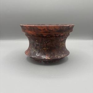 May include: A decorative, handcrafted ceramic pot with a unique hourglass shape. The pot features a textured surface with a blend of reddish-brown and gold tones. The rim has a slightly uneven edge, adding to its rustic charm.