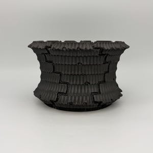 May include: A matte black planter with a wide, flared rim and a cylindrical body. The exterior surface features a repeating vertical ribbed texture interrupted by jagged, puzzle-like seams that run horizontally and vertically.
