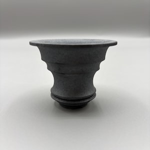 May include: A dark gray, decorative ceramic vessel with a flared rim and a tiered, pedestal base. The object has a textured surface and a symmetrical design, suitable for home decor or as a unique accent piece.