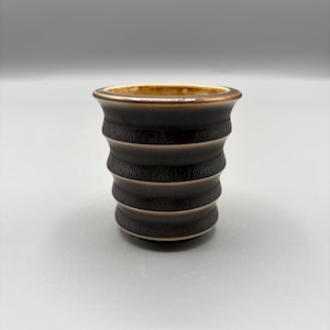 May include: A small, dark brown ceramic cup with a textured, ridged design. The rim and interior have a lighter brown color. The cup is cylindrical in shape and sits on a plain, light gray background.