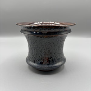 May include: A small, flared ceramic cup with a dark, textured, hammered-style surface. The exterior features a deep charcoal and black glaze with subtle metallic brown undertones, while the wide, smooth rim transitions into a rich, glossy bronze.