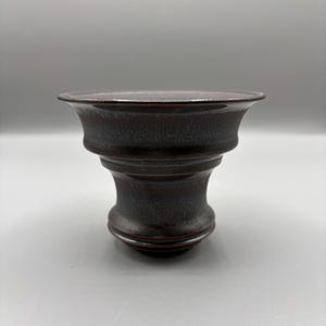 May include: A dark brown ceramic bowl with a flared rim and a pedestal base. The bowl has a textured surface and a glossy finish. The design is simple and elegant, suitable for serving or display.