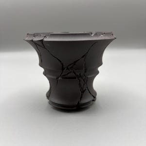 May include: A dark charcoal gray ceramic vessel with a flared rim and a tapered base. The exterior surface features a distinct pattern of deep, jagged cracks running vertically and horizontally across the body, creating a textured, fragmented appearance.