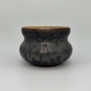 May include: A ceramic bowl with a rounded, bulbous body and a wide, open mouth. The exterior features a reactive glaze with a blend of dark charcoal, deep blue, and brown tones, creating a textured, mottled appearance across the surface.