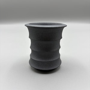 May include: A dark gray, textured ceramic vase with a flared top and a series of horizontal, ridged bands around the body. The vase has a cylindrical shape and sits on a light gray surface.