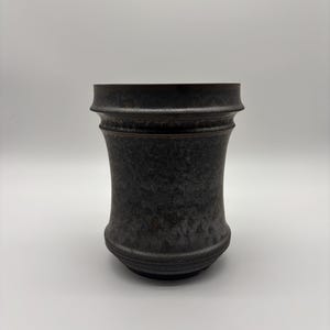 May include: A dark gray ceramic cup with a textured, mottled finish. The vessel features a wide, flared rim, a curved body that tapers inward toward the center, and a rounded base with subtle horizontal ridges near the bottom edge.