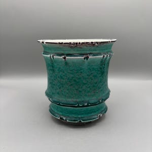 May include: A cylindrical ceramic planter featuring a vibrant teal and green mottled glaze with a textured, dimpled surface. The vessel has a flared rim and a slightly wider base, both accented with dark, distressed edges that highlight the form.