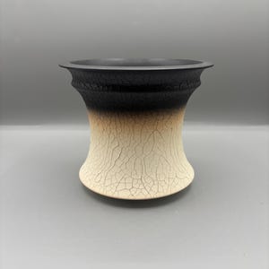 May include: A ceramic vase with a unique design. The top is black, transitioning to a tan middle, and ending in a cracked white base. The vase has a flared rim and a bulbous shape, suitable for home decor.