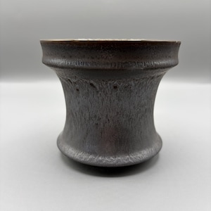 May include: A dark, metallic-toned ceramic cup with a flared rim and a narrow, concave waist. The exterior surface features a textured, mottled finish with subtle variations in color, transitioning from deep charcoal to lighter gray tones.