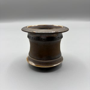 May include: A handcrafted, brown ceramic vessel with a wide, flat rim and a rounded base. The vessel has a cylindrical body with a slight hourglass shape. The rim and base have a lighter, metallic glaze, creating a contrast with the darker brown body.