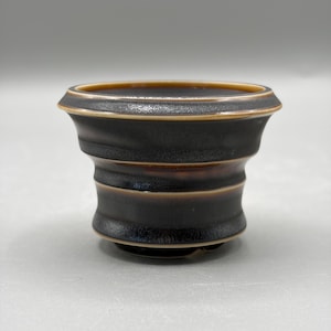 May include: A small, dark brown ceramic cup with a wide, flared rim and a tapered body. The surface features a textured, matte finish with subtle horizontal ridges and a lighter brown glaze accenting the rim and the base of the cup.