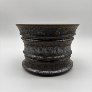 May include: A ceramic bowl with a flared rim and a tapered waist, featuring a dark, metallic-toned glaze with mottled black, charcoal, and bronze-coloured textures. The surface has a distinct horizontal ribbed pattern around the centre.