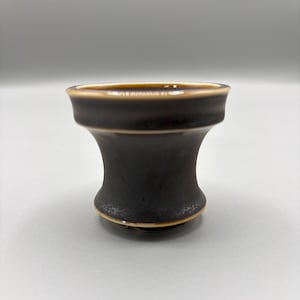 May include: A small, flared ceramic bowl with a dark brown, matte finish and a smooth, glossy tan rim. The vessel features a concave, hourglass-shaped body that tapers toward the base, with a subtle speckled texture visible near the bottom edge.