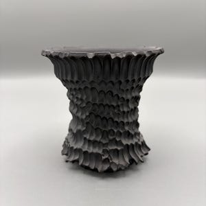 Puede incluir: Un jarrón decorativo gris oscuro texturizado con un patrón tallado único. El jarrón tiene una parte superior ancha y acanalada y un cuerpo bulboso y texturizado. La superficie está cubierta con un diseño repetitivo en forma de escamas, dándole una apariencia táctil.