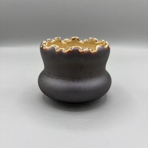 May include: A small, rounded ceramic vessel with a dark, matte charcoal-colored exterior and a textured, speckled finish. The rim features a distinct, wavy, scalloped edge with a contrasting light beige glaze that drips down into the interior.
