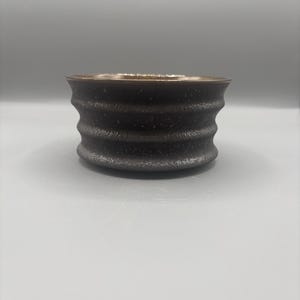 May include: A small, dark brown ceramic bowl with a textured, ridged exterior. The rim has a lighter, golden-brown color. The bowl is cylindrical with a slightly flared top. The surface has a speckled appearance.