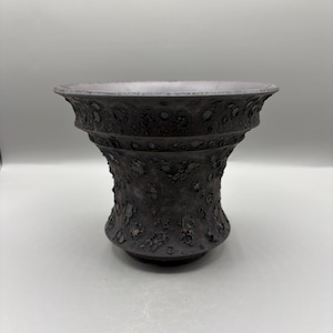 May include: A dark brown ceramic vase with a textured, pitted surface. The vase has a wide, flared rim and a bulbous body, with a textured pattern. The vase is likely for decorative purposes.