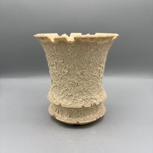 May include: A beige ceramic vessel with a flared rim and a textured, porous surface. The exterior features a complex, cratered pattern with small holes and raised edges, narrowing towards the base which has a distinct, tiered circular foot.