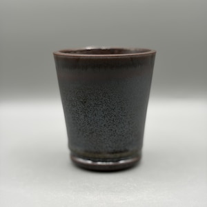 Könnte beinhalten: Ein Keramikbecher mit konischer zylindrischer Form und einer dunklen, gesprenkelten schwarz-braunen Glasur. Die Oberfläche weist ein strukturiertes, punktiertes Finish auf, das in einen glatten, dunkleren Rand übergeht, mit einem leicht ausgestellten Boden.