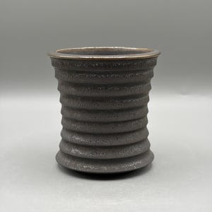 Könnte beinhalten: Ein dunkel anthrazitfarbener Keramikbecher mit einer texturierten, gerippten Außenseite mit horizontalen Rillen. Das Gefäß hat einen leicht ausgestellten Rand und einen metallisch wirkenden Innenrand sowie eine matte, gesprenkelte Oberfläche.
