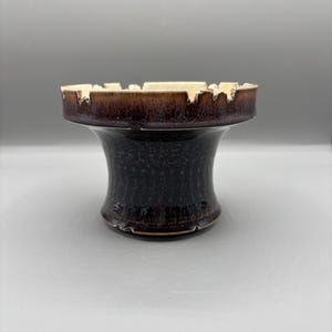 May include: A ceramic ashtray with a dark brown, speckled body and a lighter brown rim. The rim has a unique, jagged edge design. The ashtray has a pedestal base and a glossy finish.
