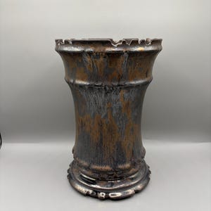May include: A decorative vase with a textured metallic finish. The vase has a cylindrical shape with a flared top and a detailed base. The color scheme includes shades of bronze, silver, and dark gray, creating a rustic aesthetic.