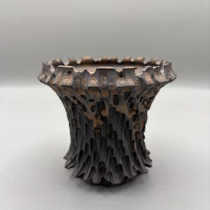 Könnte beinhalten: Eine handgefertigte, strukturierte Vase mit dunkelbraunem Äußeren und bronzefarbenem Inneren. Die Vase hat ein einzigartiges, organisches Design mit einem gerüschten Rand und einer rauen, geschnitzten Oberfläche. Die Vase ist etwa 15 cm hoch.