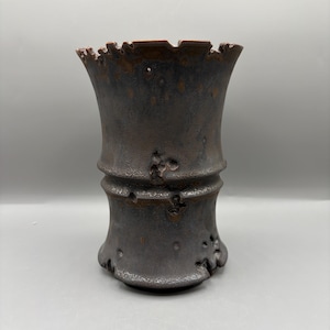 Pode incluir: Um vaso de cerâmica cinza escuro com uma superfície texturizada e uma borda única e ondulada. O vaso tem uma forma cilíndrica com um topo ligeiramente alargado e cristas horizontais. A borda interna é de cor marrom avermelhado.