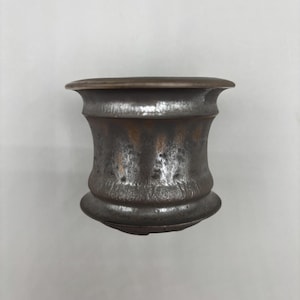 May include: A weathered, dark metal mortar with a flared rim and a rounded base. The mortar has a cylindrical body with a slightly hourglass shape. The metal has a mottled, aged appearance, suggesting it is antique or vintage.