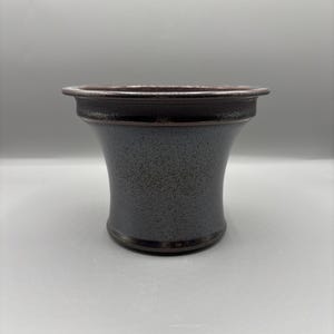 May include: A ceramic vase with a flared shape and a speckled, gray-blue glaze. The rim and base feature a dark brown band. The vase has a slightly textured surface and a simple, elegant design, suitable for various decorative purposes.