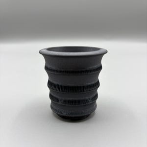 May include: A dark gray ceramic vase with a textured, ribbed design. The vase has a flared rim and a cylindrical body with horizontal bands of detailed texture. The vase is set against a plain, light background.