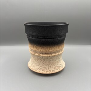 Könnte beinhalten: Eine handgefertigte Keramikvase mit einer einzigartigen, strukturierten Oberfläche. Der obere Teil ist schwarz, geht in Beige über und endet in einem cremefarbenen Sockel. Die Vase hat eine zylindrische Form mit einem ausgestellten Rand und einem rissigen, gealterten Aussehen.
