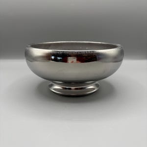 May include: A silver-colored bowl with a rounded body and a pedestal base. The bowl has a smooth, reflective surface and a wide opening. The pedestal base is circular and provides stability. The bowl is likely for serving or display.