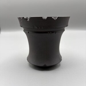 May include: A dark gray, ceramic vase with a unique, hourglass-like shape. The top edge has a jagged, distressed design. The vase is a solid color and sits on a white surface. The design is modern and minimalist.