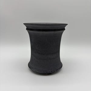May include: A matte black ceramic vessel with a flared rim and a tapered, concave body. The surface features a subtle, mottled texture with variations in tone, creating a dark, monochromatic appearance with a defined horizontal ridge near the top.