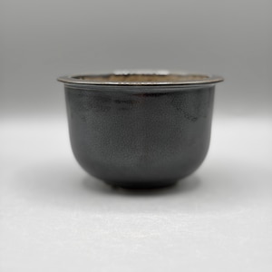 Peut inclure: Un petit bol en céramique arrondi avec un vernis gris anthracite à l'aspect métallique. La surface extérieure présente une texture fine et subtile, tandis que le bord est légèrement évasé et doté d'une bordure sombre et lisse.