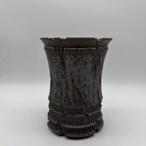 May include: A dark brown ceramic vase with a textured, bark-like surface and a flared, notched rim. The lower section features a repeating vertical ribbed pattern, while the upper body displays a mottled, weathered finish with deep grooves.