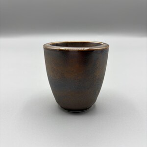 May include: A small, handcrafted ceramic cup with a dark brown exterior and a gold-colored rim. The cup has a smooth, tapered shape and a slightly textured surface. The interior is a darker shade of brown.