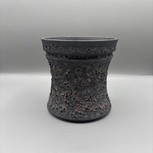 Puede incluir: Un jarrón de cerámica gris oscuro con una superficie texturizada y rugosa. El jarrón tiene una base ancha que se estrecha hacia adentro y luego se ensancha en la parte superior. La textura tiene pequeños agujeros irregulares.
