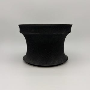 May include: A matte black planter with a concave, hourglass-shaped silhouette. The surface features a subtle, fine-grained texture throughout the exterior. The vessel has a wide, flared rim at the top and a matching flared base, creating a symmetrical form.