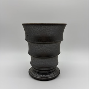 May include: A dark charcoal gray ceramic cup with a flared rim and a tiered, stepped silhouette. The surface features a fine, speckled texture throughout, with a wider opening at the top that tapers down to a circular base with a defined foot.