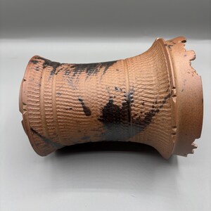 May include: A brown ceramic vase with a textured surface and black paint details. The vase has a cylindrical shape with a flared top and bottom. The surface has a woven pattern.