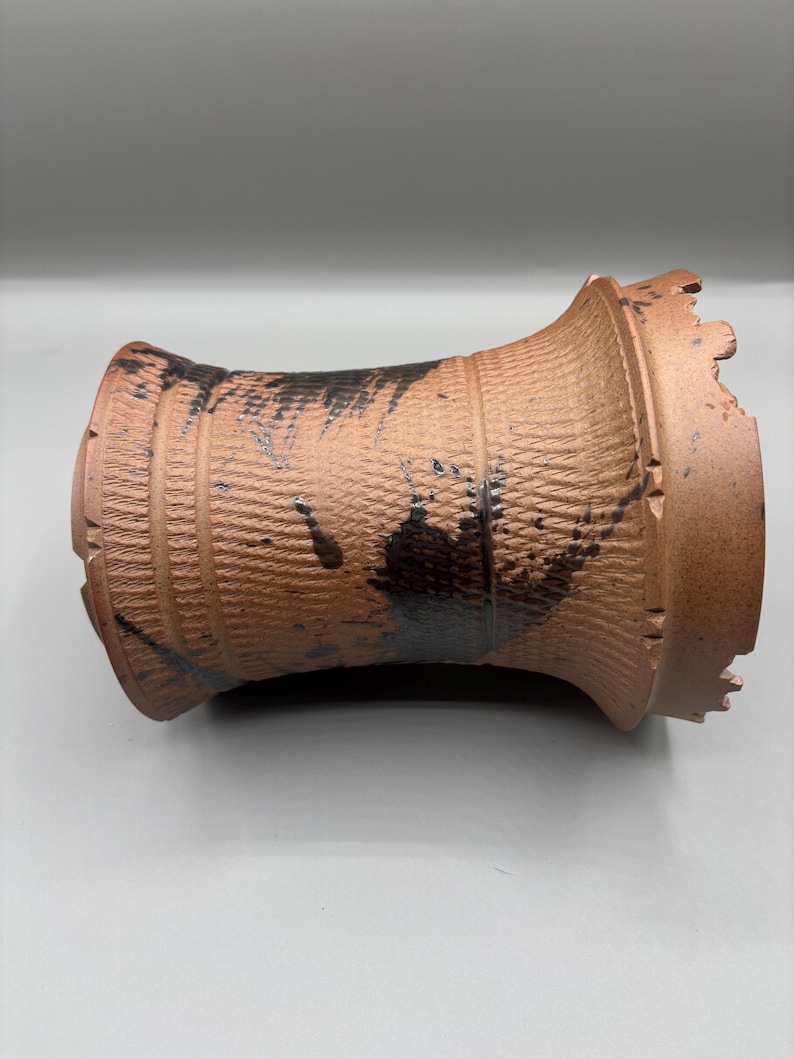 May include: A brown ceramic vase with a textured surface and black paint details. The vase has a cylindrical shape with a flared top and bottom. The surface has a woven pattern.