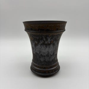 May include: A ceramic vase with a flared rim and a tapered body. The surface features a dark, mottled glaze with a textured, metallic-looking finish that transitions from deep black at the base to a brownish-bronze hue near the top rim.