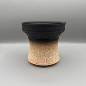 May include: A handcrafted ceramic pot with a unique design. The top is black, transitioning to a tan base with a cracked, textured finish. The pot has a wide, open top and a flared, rounded shape.