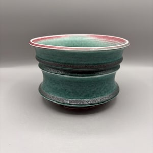 May include: A ceramic bowl with a flared rim, featuring a mottled teal and green glaze. The exterior has two distinct horizontal ridges with a dark, textured finish. The interior rim shows a thin band of pinkish-red color against the green glaze.